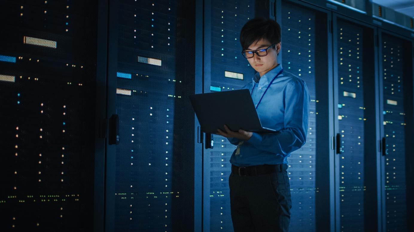 Blue-shirted man working on black laptop in front of a supercomputer.