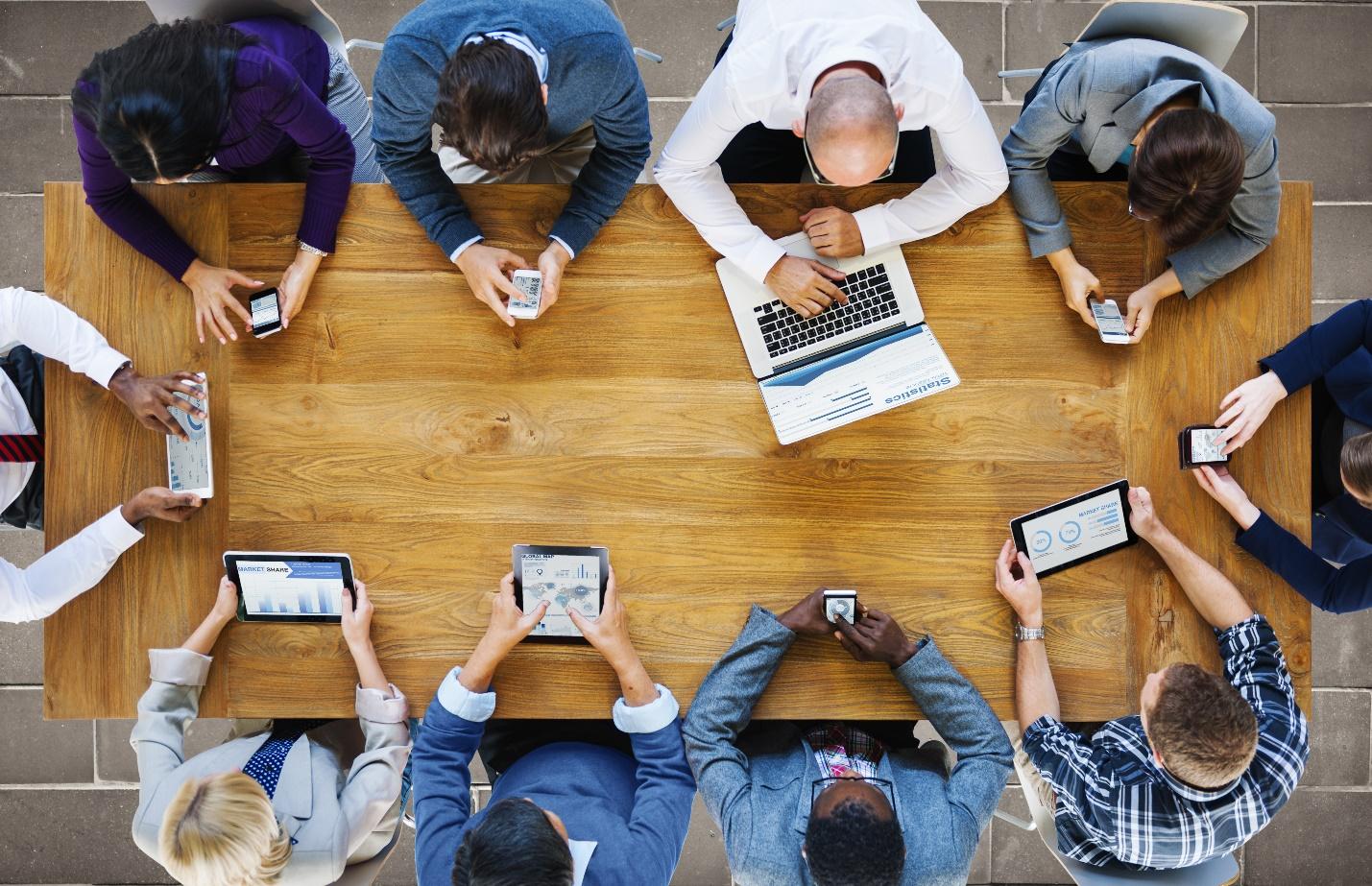 An overhead photo of business professionals around a conference table using notepads, phones, and laptops to work.