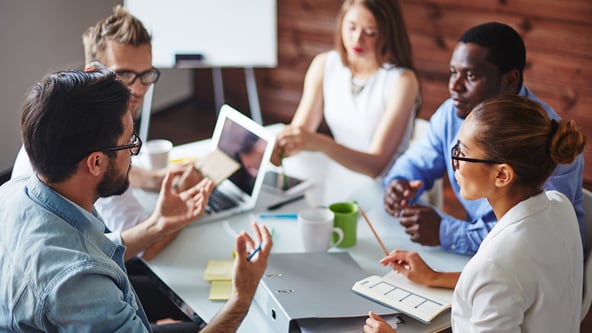 Five people are sitting around a table talking. There two women and three men. They look like they are in a meeting. 