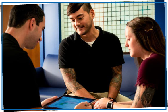 Three people talking over a table that has ConnectWise Manage on it. There are two men in black polos, one has tattoos. There is also a woman wearing pink with tattoos as well. 