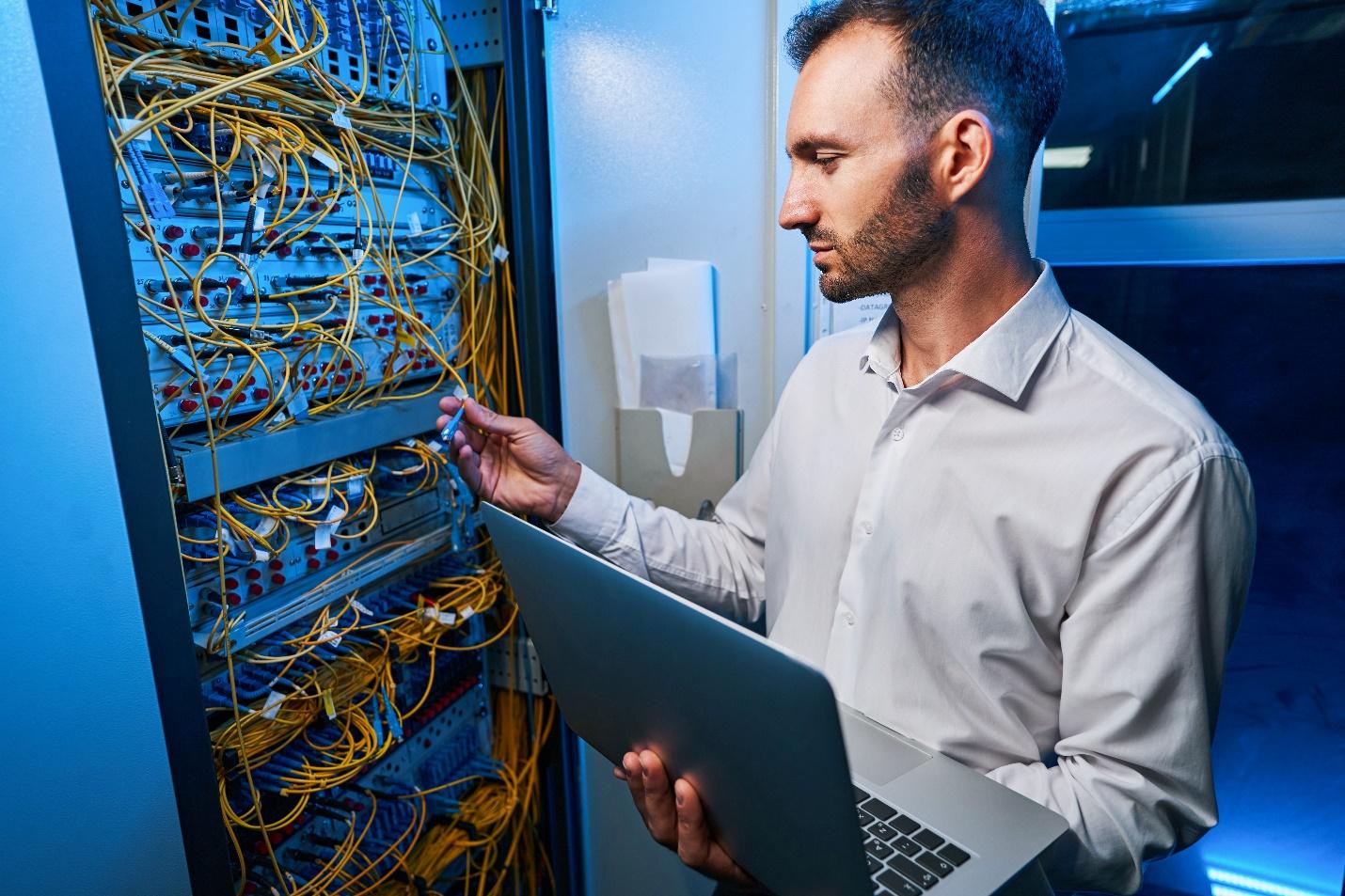 A male IT technician holds a laptop while connecting a cable to a server rack.